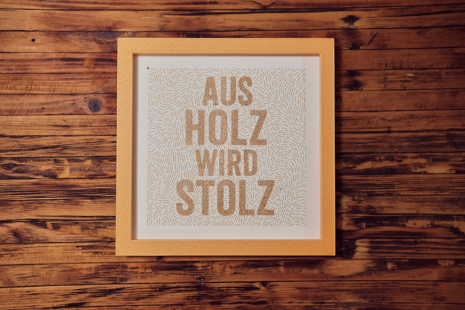 Gerahmtes Plotter-Kunstwerk 'Aus Holz wird Stolz' in Goldton mit Strichtextur auf Holzhintergrund